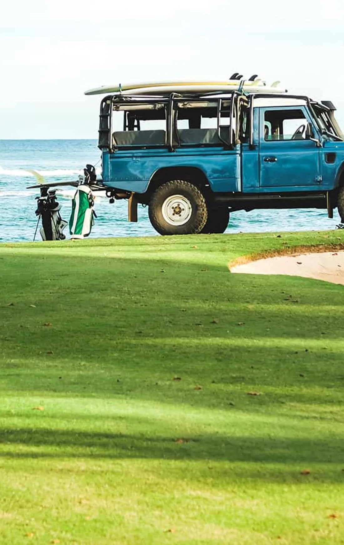 A blue Land Rover Defender with surfboards on the roof, parked above the ocean on a golf course, a golf bag leaning against it